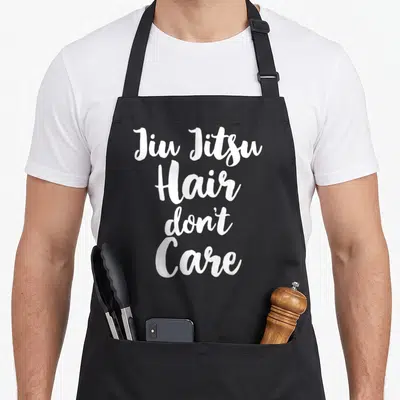 Jiu christmas apron - White stacked script text reading Jiu Jitsu Hair Don't Care — shown on a black cotton canvas apron against a white background