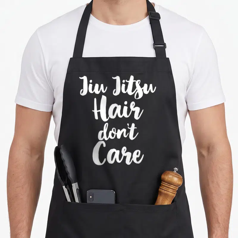 Jiu christmas apron - White stacked script text reading Jiu Jitsu Hair Don't Care — shown on a black cotton canvas apron against a white background