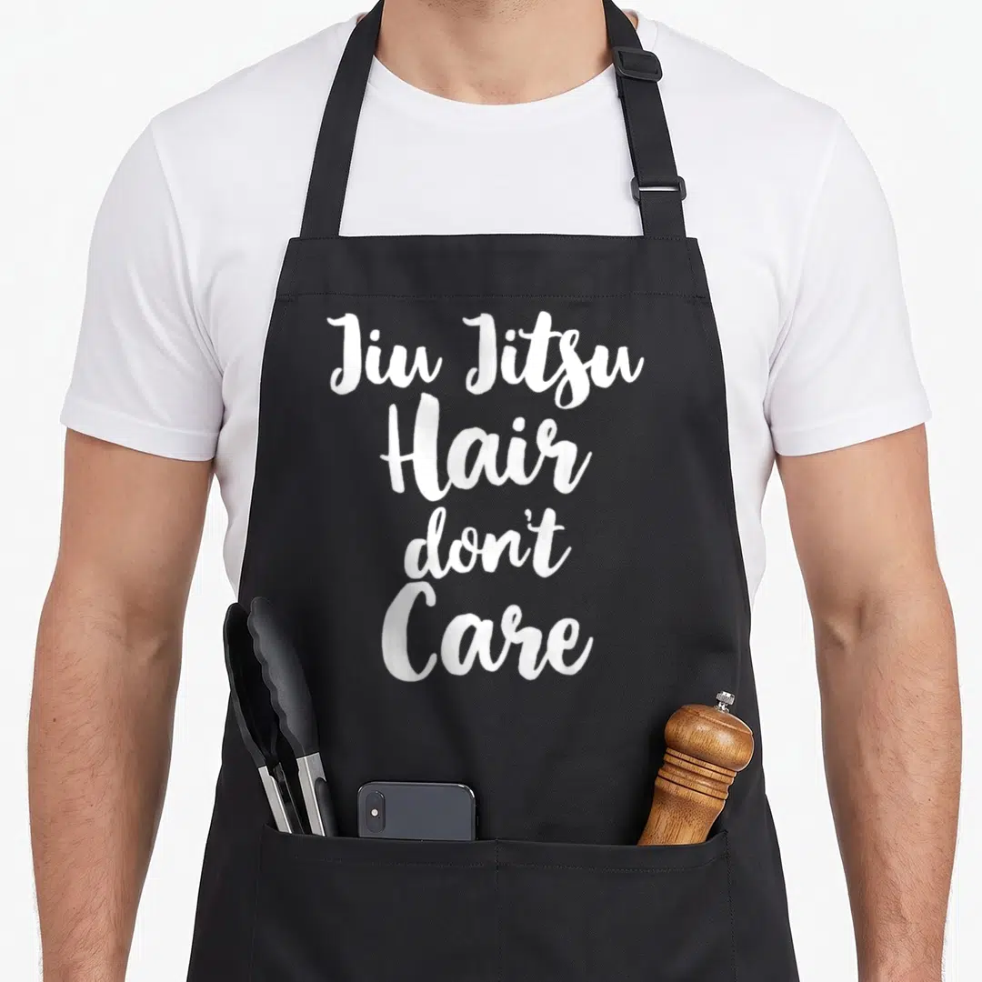 Jiu christmas apron - White stacked script text reading Jiu Jitsu Hair Don't Care — shown on a black cotton canvas apron against a white background