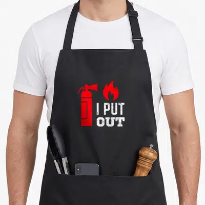 Funny fireman apron - Red fire extinguisher and flame next to stacked white text I PUT OUT — shown on a black cotton canvas apron against a white background