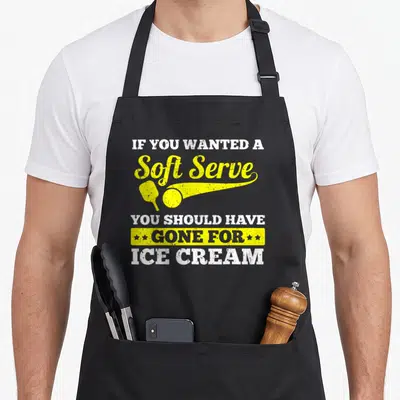 Sport to serve man apron - Pickleball paddle and ball graphic with distressed white and yellow text reading soft serve — shown on a black cotton canvas apron against a white background