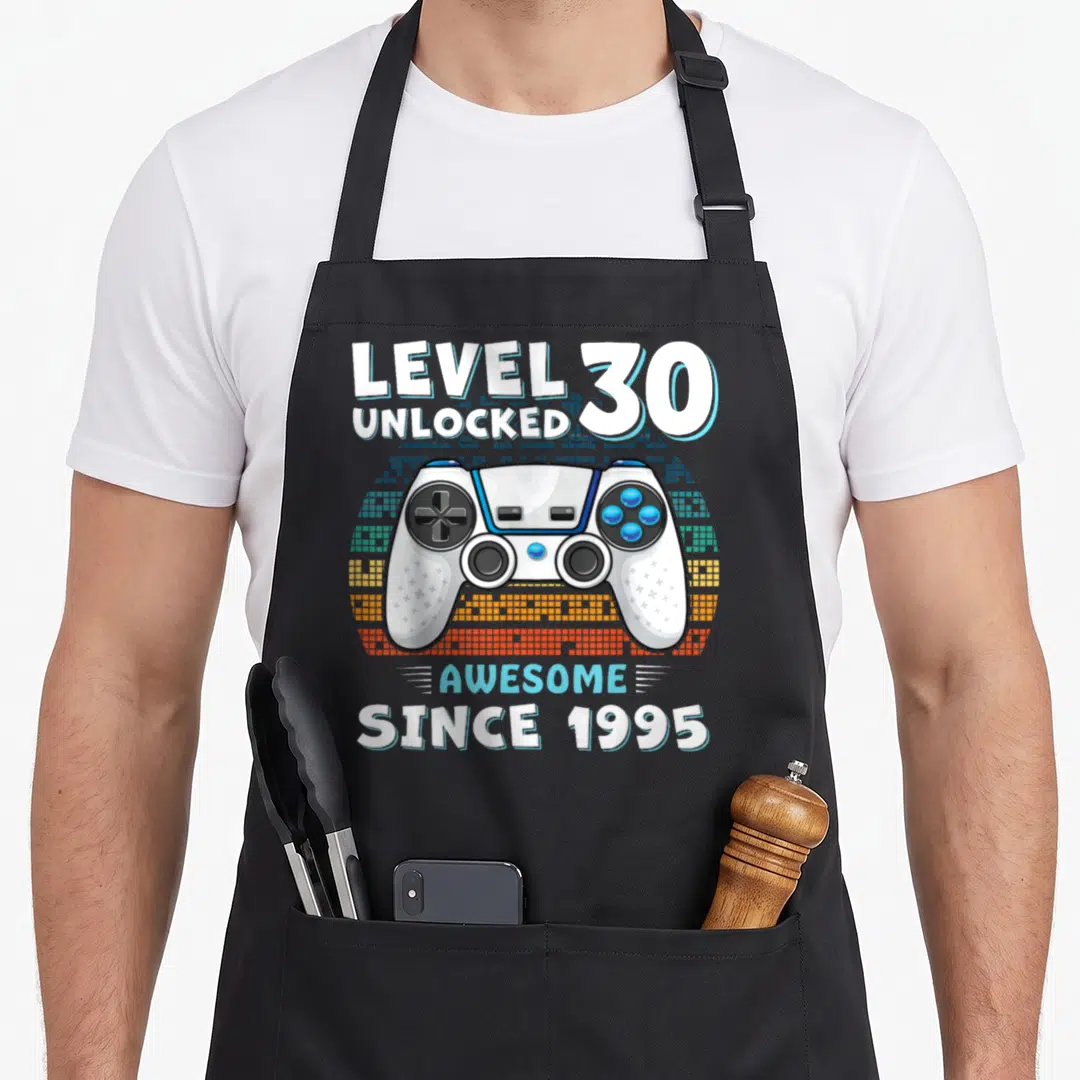 30yr gag aprons - Game controller with LEVEL 30 UNLOCKED and AWESOME SINCE 1995 text over pixelated semi-circle — shown on a black cotton canvas apron against a white background