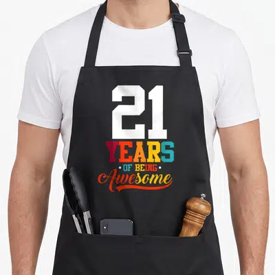 21st born day apron - Stacked colorful text 21 YEARS OF BEING Awesome in white red orange yellow blue — shown on a black cotton canvas apron against a white background
