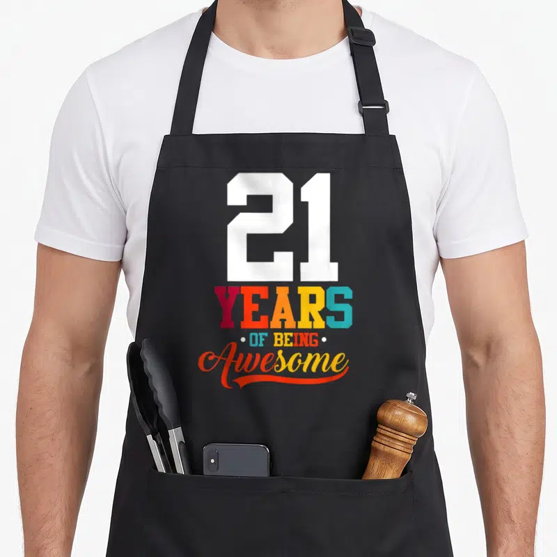 21st born day apron - Stacked colorful text 21 YEARS OF BEING Awesome in white red orange yellow blue — shown on a black cotton canvas apron against a white background