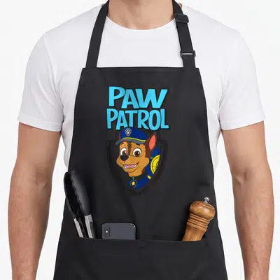Badge chase birthday apron - Cartoon brown dog in blue police hat inside grey shield below stacked blue text — shown on a black cotton canvas apron against a white background