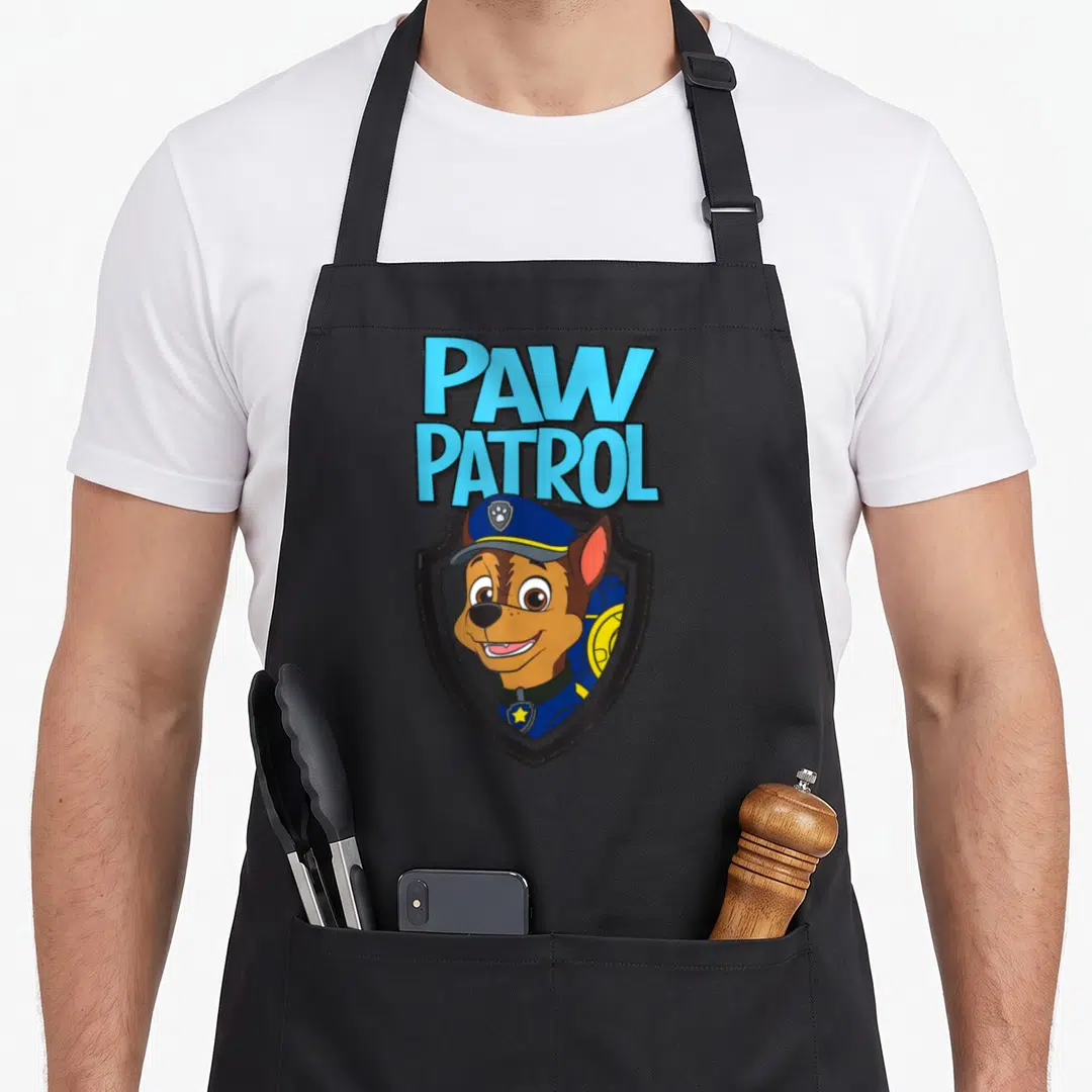Badge chase birthday apron - Cartoon brown dog in blue police hat inside grey shield below stacked blue text — shown on a black cotton canvas apron against a white background