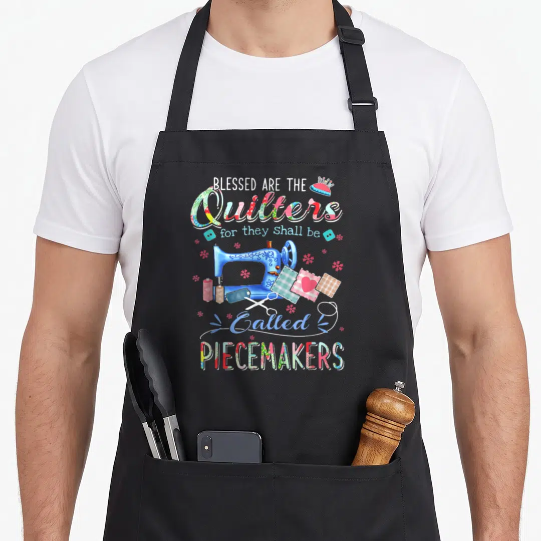Blessed quilter apron - Blue sewing machine surrounded by fabric squares and text Blessed Are The Quilters Called — shown on a black cotton canvas apron against a white background