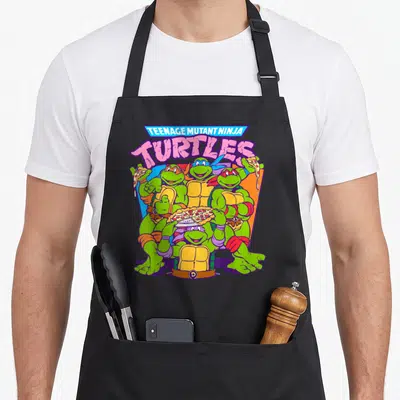 Smile pizza apron - Stacked TMNT text above four turtles holding pizza slices and a whole pie — shown on a black cotton canvas apron against a white background