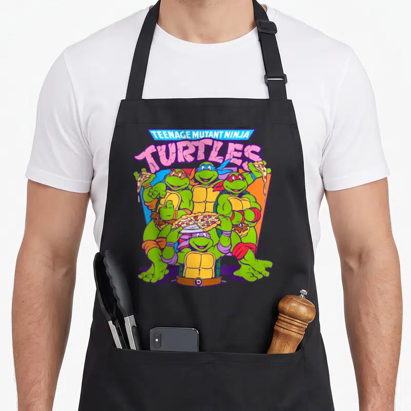 Smile pizza apron - Stacked TMNT text above four turtles holding pizza slices and a whole pie — shown on a black cotton canvas apron against a white background