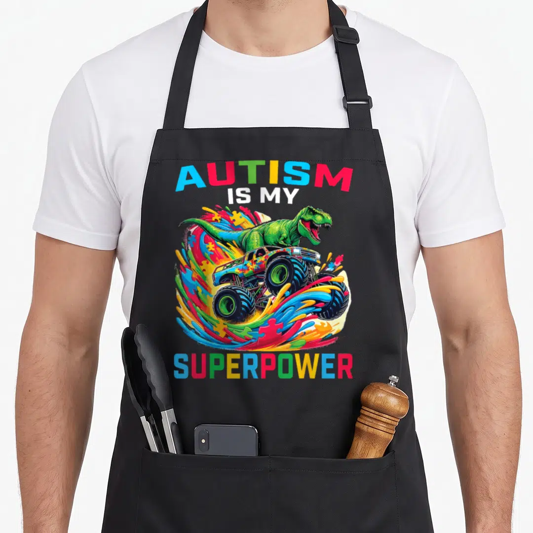 Autism truck apron - Monster truck with T-Rex and puzzle pieces under AUTISM IS MY SUPERPOWER text — shown on a black cotton canvas apron against a white background