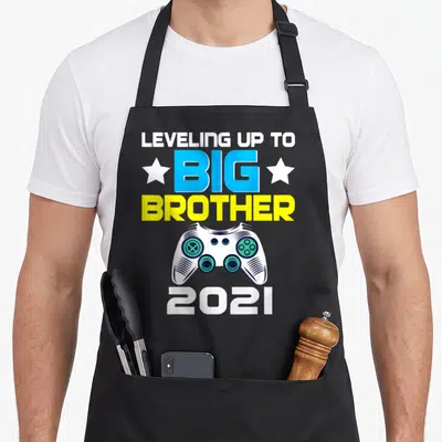 Announcement brother apron - Game controller graphic with LEVELING UP TO BIG BROTHER 2021 text and stars — shown on a black cotton canvas apron against a white background
