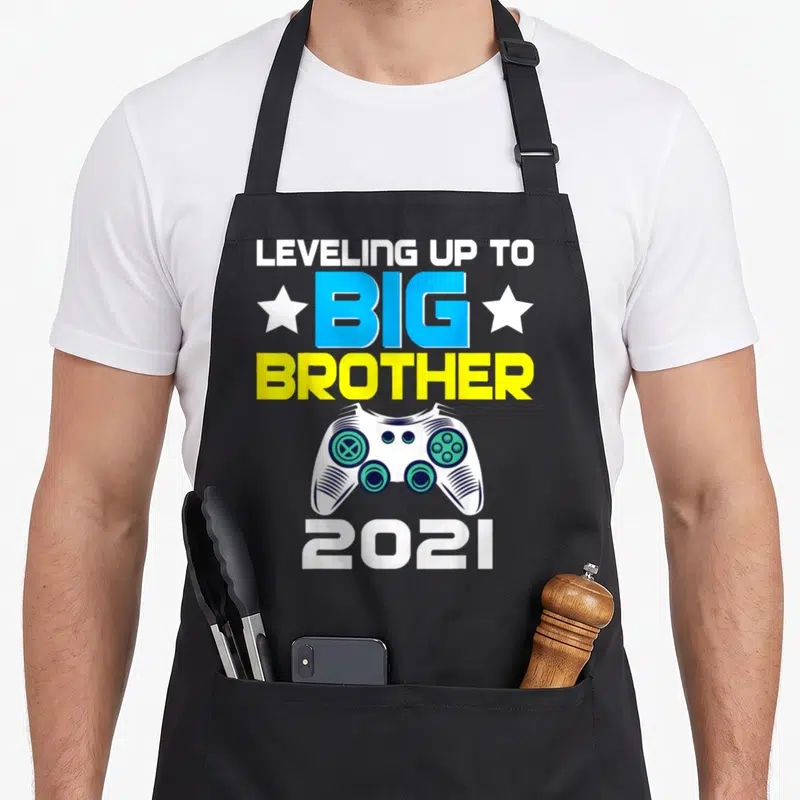 Announcement brother apron - Game controller graphic with LEVELING UP TO BIG BROTHER 2021 text and stars — shown on a black cotton canvas apron against a white background