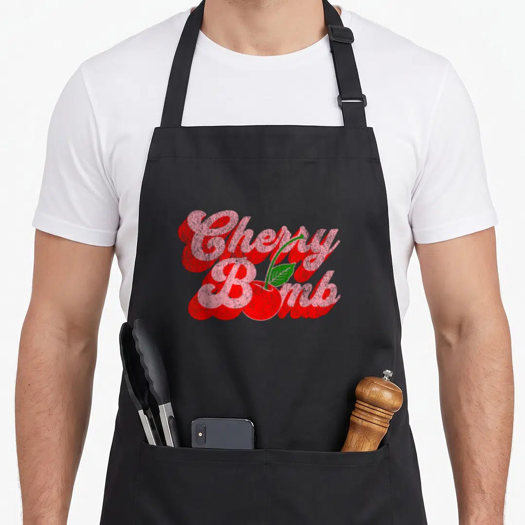 Bomb 70s cherry apron - Stacked pink and red Cherry Bomb text with a red cherry and green leaf graphic — shown on a black cotton canvas apron against a white background