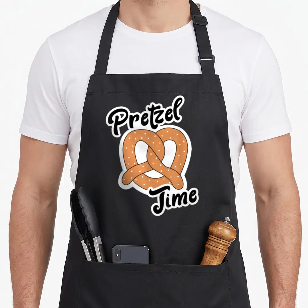 Cute pretzels apron - Pretzel Time text above and below a brown pretzel with white salt specks and thick white border — shown on a black cotton canvas apron against a white background