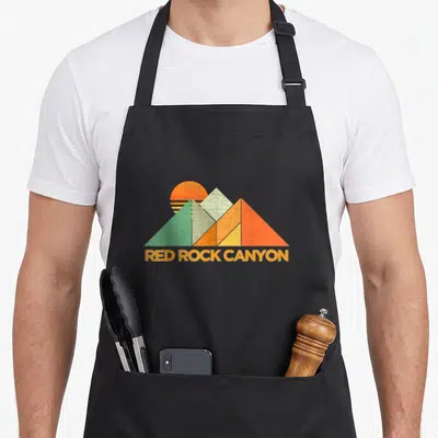 Cute red rock canyon apron - Retro geometric mountains in orange, yellow, green with RED ROCK CANYON text below — shown on a black cotton canvas apron against a white background