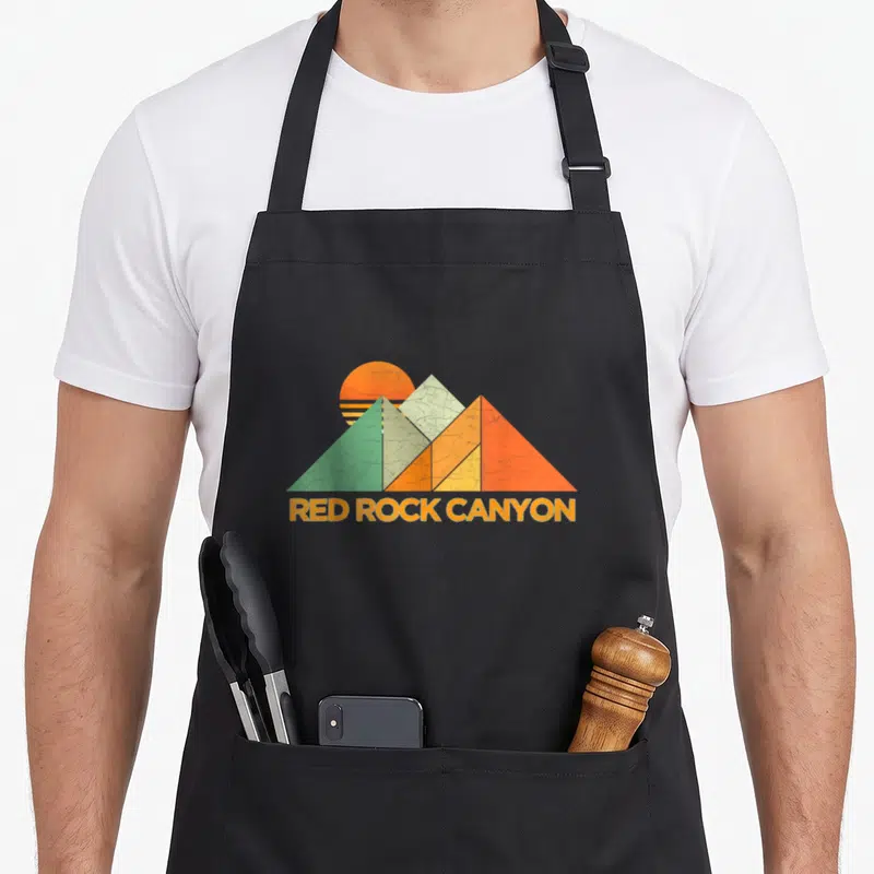 Cute red rock canyon apron - Retro geometric mountains in orange, yellow, green with RED ROCK CANYON text below — shown on a black cotton canvas apron against a white background