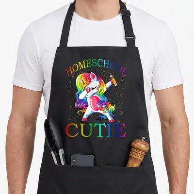 School homeschool unicorn apron - Cartoon white unicorn dabbing with rainbow mane holding yellow pencil surrounded — shown on a black cotton canvas apron against a white background