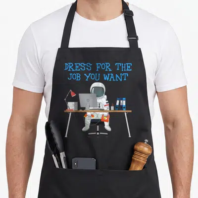 Dress cosmonaut apron - Cartoon astronaut in white suit at brown desk holding orange phone under blue text — shown on a black cotton canvas apron against a white background
