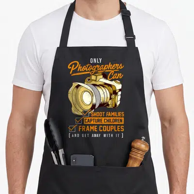 Pun photographer apron - Camera illustration with checklist text reading shoot families capture children frame couples — shown on a black cotton canvas apron against a white background