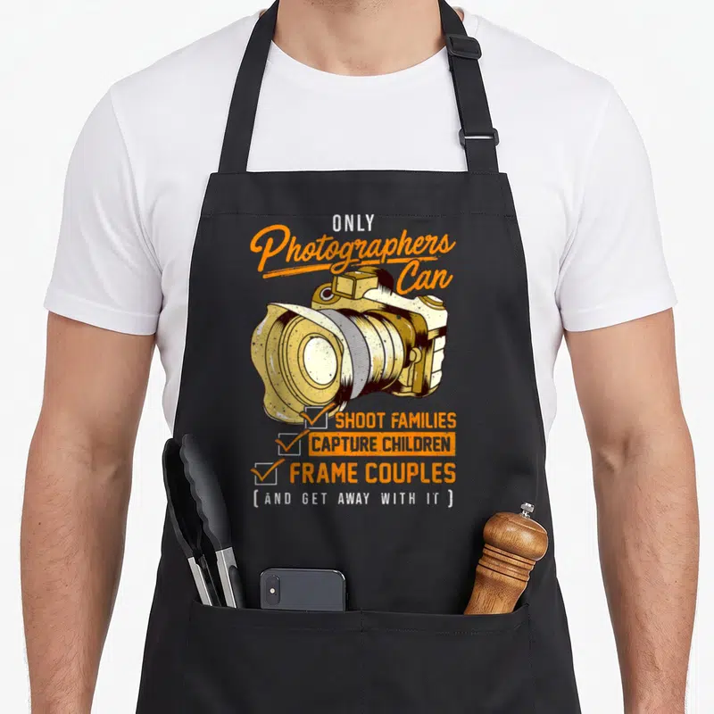 Pun photographer apron - Camera illustration with checklist text reading shoot families capture children frame couples — shown on a black cotton canvas apron against a white background