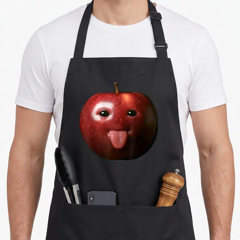 Silly meme apron - Red apple with two eyes and pink tongue sticking out on black background — shown on a black cotton canvas apron against a white background