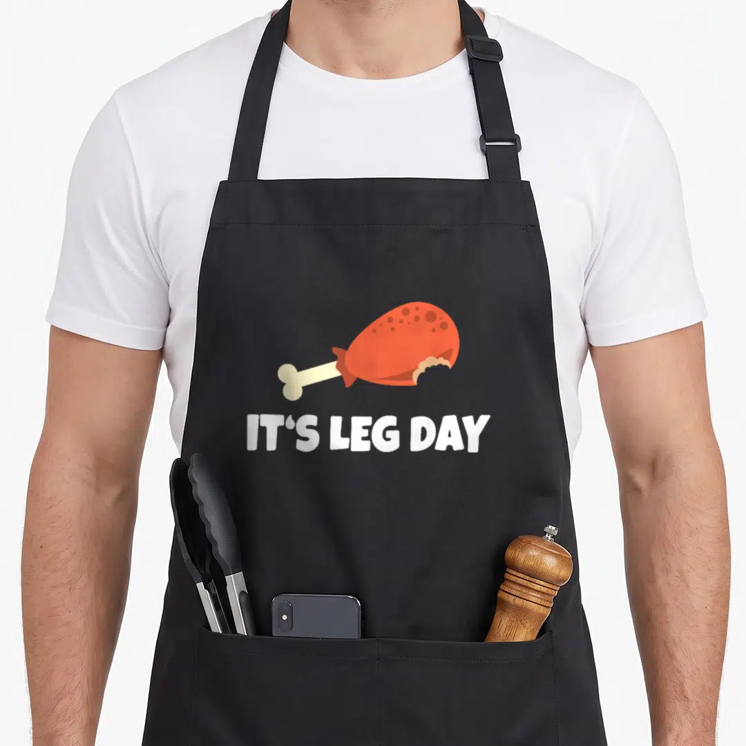 Leg workout thanksgiving day aprons - Cartoon orange chicken leg above white It's Leg Day text on black — shown on a black cotton canvas apron against a white background