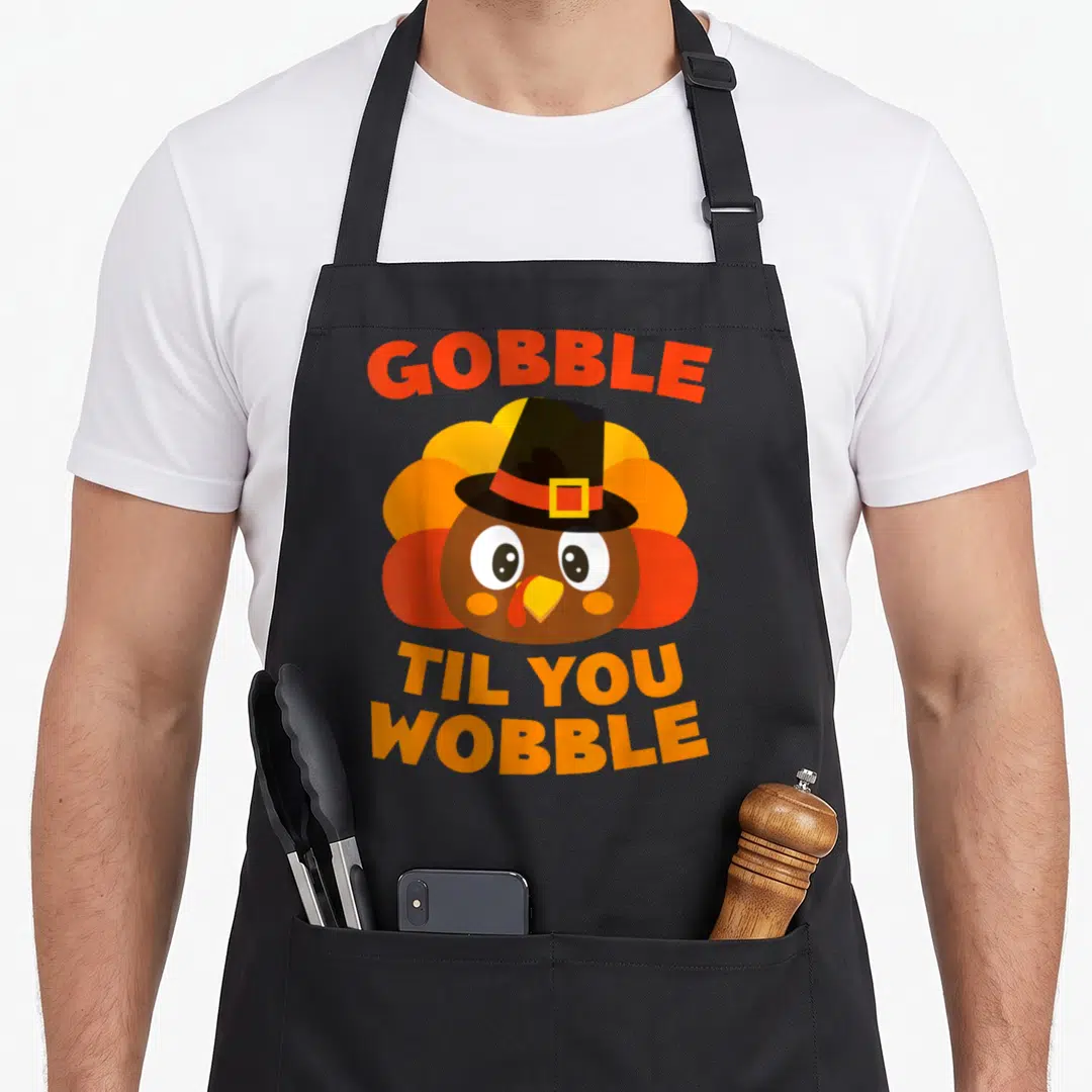 Gobble thanksgiving day aprons - Cartoon turkey head in orange and brown with black pilgrim hat and text GOBBLE TIL YOU — shown on a black cotton canvas apron against a white background