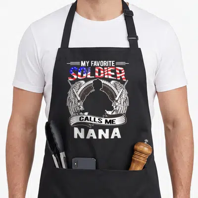 Proud army grandma apron - Soldier silhouette with wings, rifles, and My Favorite Soldier Calls Me Nana text — shown on a black cotton canvas apron against a white background