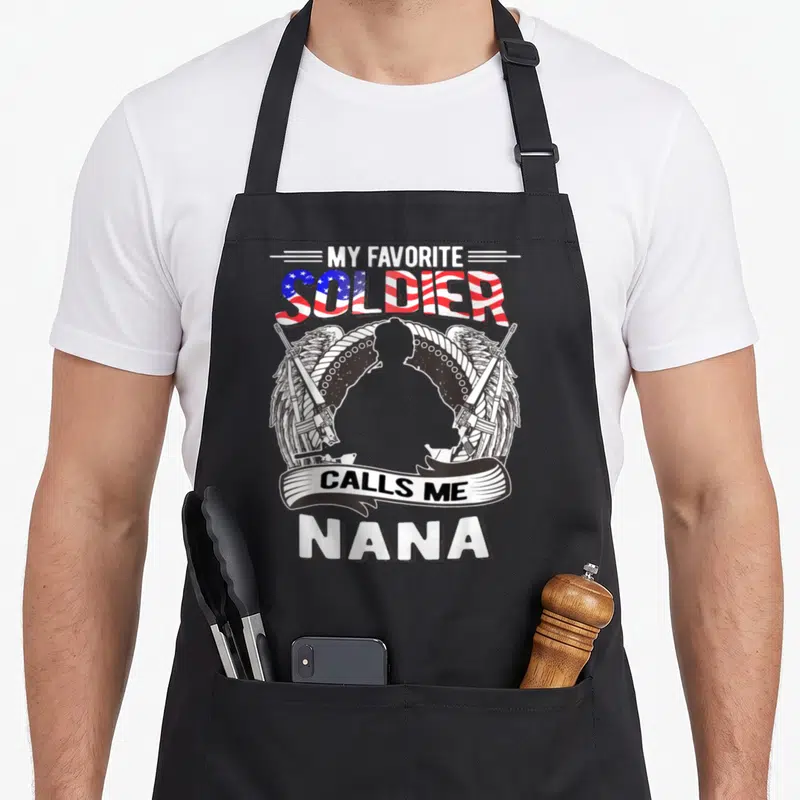 Proud army grandma apron - Soldier silhouette with wings, rifles, and My Favorite Soldier Calls Me Nana text — shown on a black cotton canvas apron against a white background