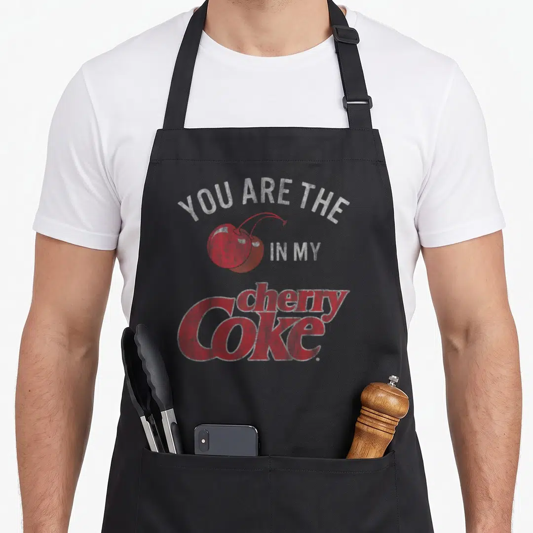 Coke cherry apron - Red and white distressed graphic of cherries with YOU ARE THE IN MY cherry Coke text — shown on a black cotton canvas apron against a white background