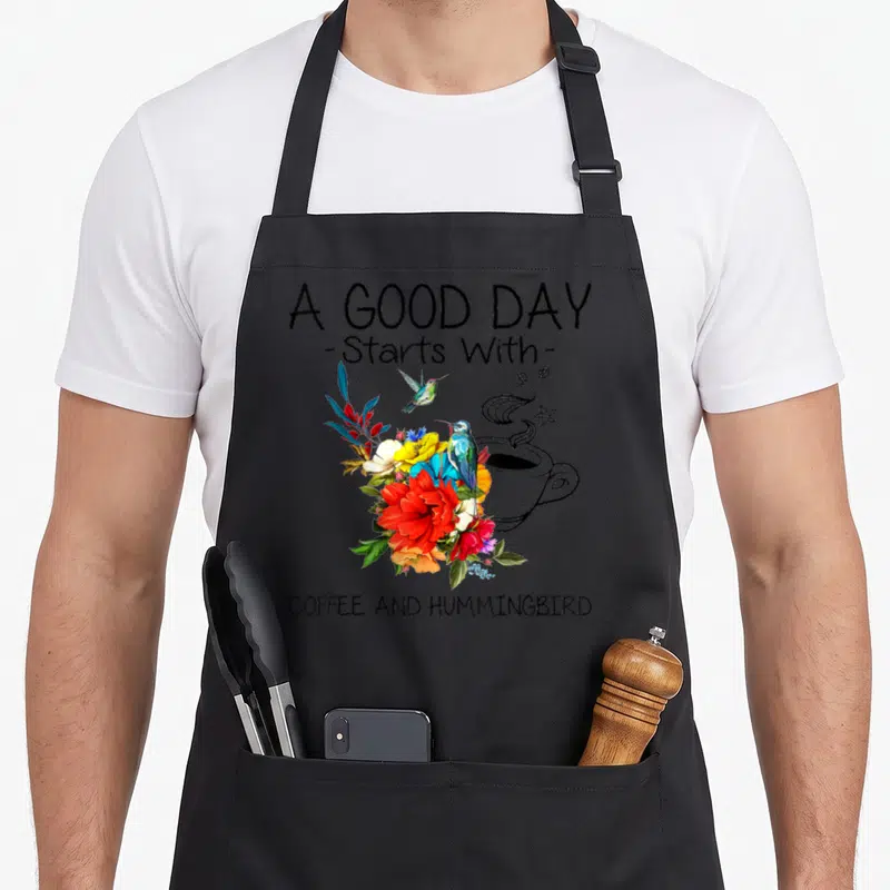 Start coffee apron - A GOOD DAY text, colorful flowers, hummingbirds, and black line-art coffee cup — shown on a black cotton canvas apron against a white background