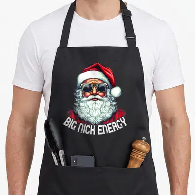 Nick santa apron - Santa Claus in sunglasses and red plaid shirt with BIG NICK ENERGY text arched below — shown on a black cotton canvas apron against a white background