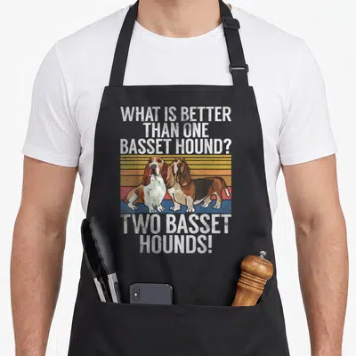 Better basset hound apron - Two basset hounds standing before retro striped background with white text — shown on a black cotton canvas apron against a white background