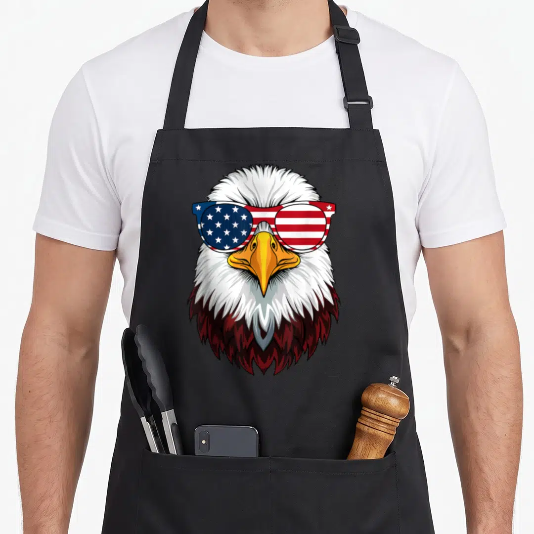 Flag patriotic eagle 4th of july apron - Bald eagle head wearing American flag sunglasses in white brown blue and red — shown on a black cotton canvas apron against a white background