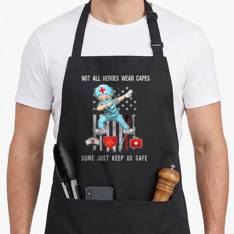 Cape nurse apron - Cartoon nurse in blue scrubs dabbing with medical icons and text about keeping us safe — shown on a black cotton canvas apron against a white background