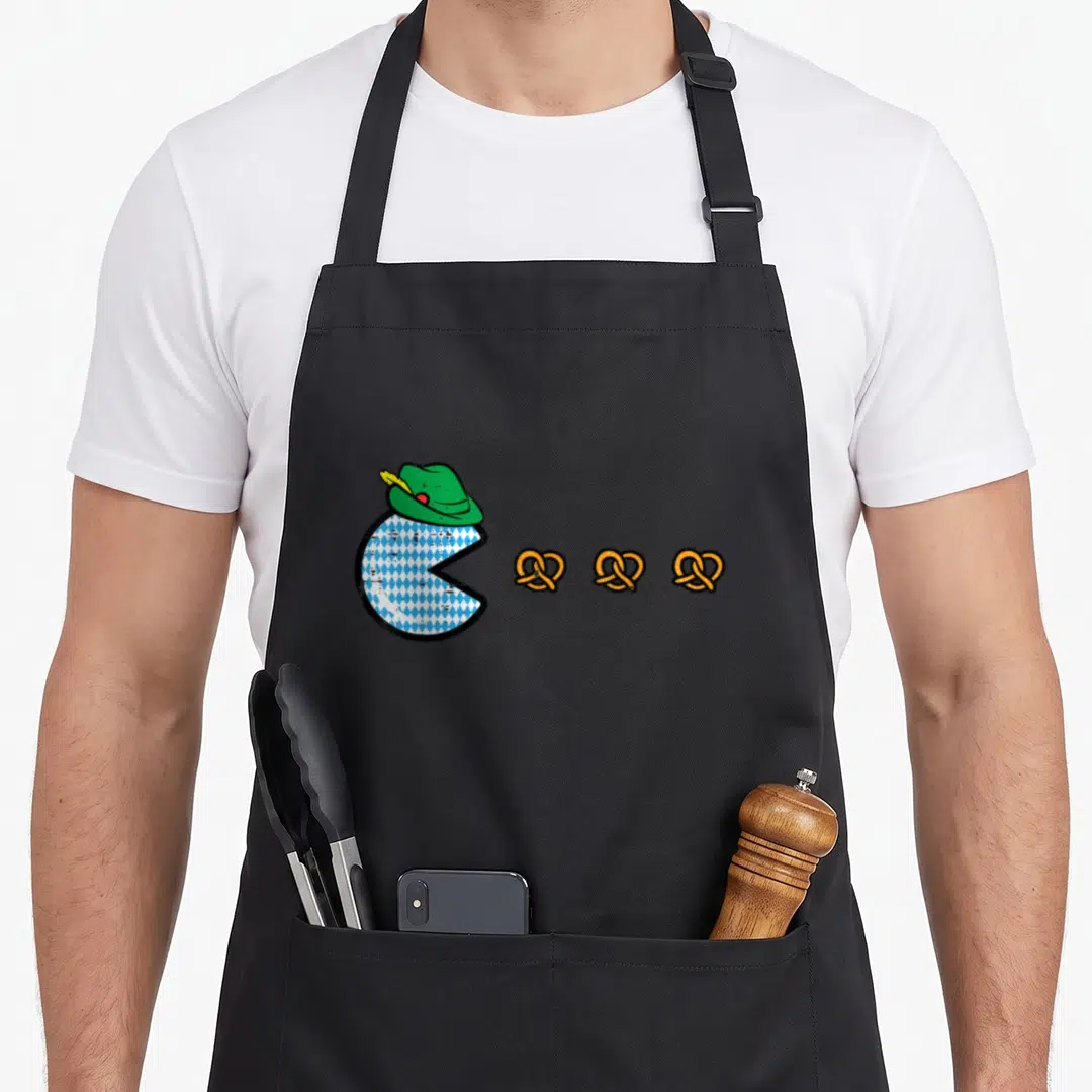 Oktoberfest video game apron - Pac-Man in blue white diamond pattern wearing green hat facing three orange pretzels — shown on a black cotton canvas apron against a white background