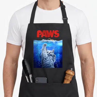 Movie cat apron - Red PAWS text above grey cat underwater reaching for swimmer in blue water — shown on a black cotton canvas apron against a white background