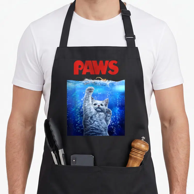 Movie cat apron - Red PAWS text above grey cat underwater reaching for swimmer in blue water — shown on a black cotton canvas apron against a white background
