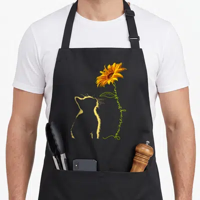Funny sunshine cat apron - Yellow cat outline looks up at sunflower with You Are My Sunshine text on lime stem — shown on a black cotton canvas apron against a white background