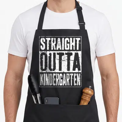 Outta kindergarten apron - White distressed text reading STRAIGHT OUTTA KINDERGARTEN inside a rectangular border — shown on a black cotton canvas apron against a white background