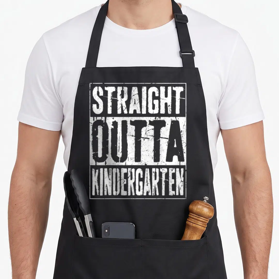 Outta kindergarten apron - White distressed text reading STRAIGHT OUTTA KINDERGARTEN inside a rectangular border — shown on a black cotton canvas apron against a white background