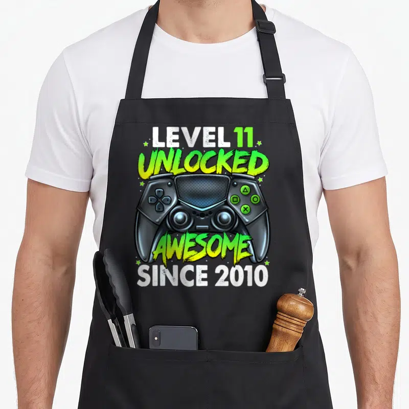 11th birthday gaming aprons - Game controller graphic with LEVEL 11 UNLOCKED AWESOME SINCE 2010 text in lime green — shown on a black cotton canvas apron against a white background