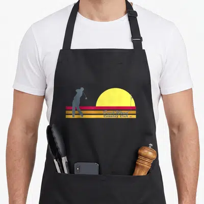 Caddyshack stripe apron - Grey golfer silhouette next to yellow sun and flag above red orange yellow stripes with blue — shown on a black cotton canvas apron against a white background