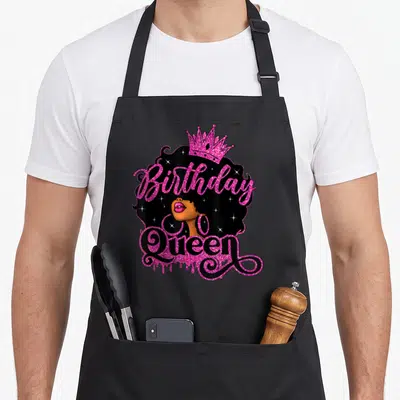 Birthday african american aprons - Cartoon woman with Birthday Queen text, pink crown, sparkles in black hair — shown on a black cotton canvas apron against a white background