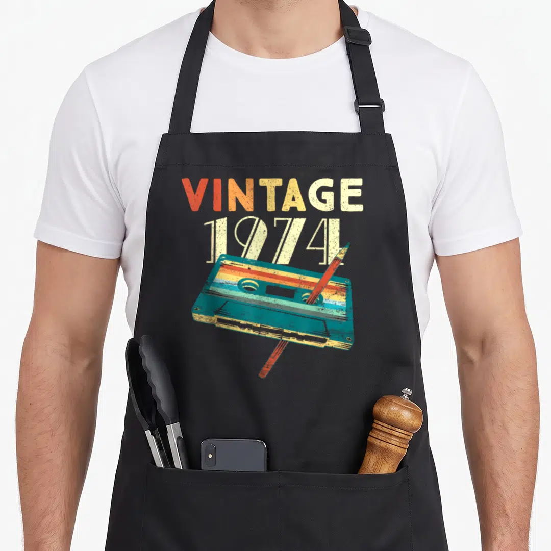 Cassette birthday apron - VINTAGE 1974 text above distressed teal cassette tape with diagonal pencil in orange cream — shown on a black cotton canvas apron against a white background