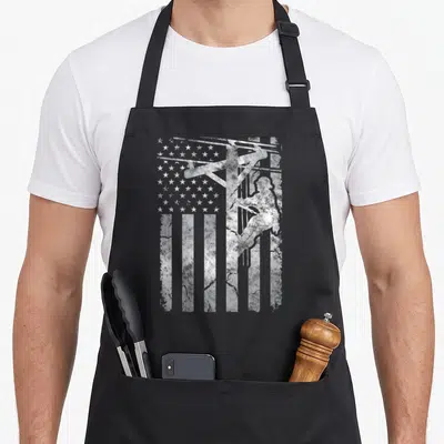 Distressed patriotic aprons - White distressed silhouette of a lineman on a utility pole overlaid on a US flag pattern — shown on a black cotton canvas apron against a white background