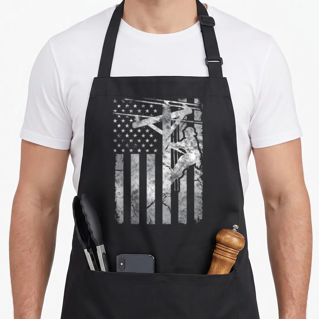 Distressed patriotic aprons - White distressed silhouette of a lineman on a utility pole overlaid on a US flag pattern — shown on a black cotton canvas apron against a white background