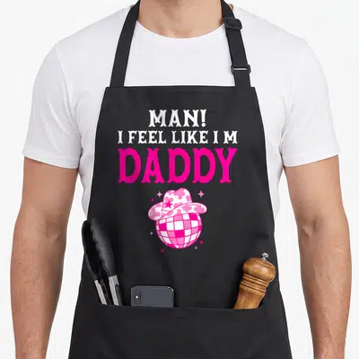 Feel cowgirl aprons - Pink disco ball wearing cowboy hat with stacked MAN I FEEL LIKE I M DADDY text — shown on a black cotton canvas apron against a white background