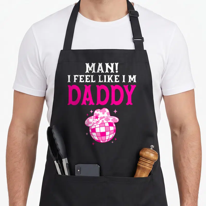 Feel cowgirl aprons - Pink disco ball wearing cowboy hat with stacked MAN I FEEL LIKE I M DADDY text — shown on a black cotton canvas apron against a white background