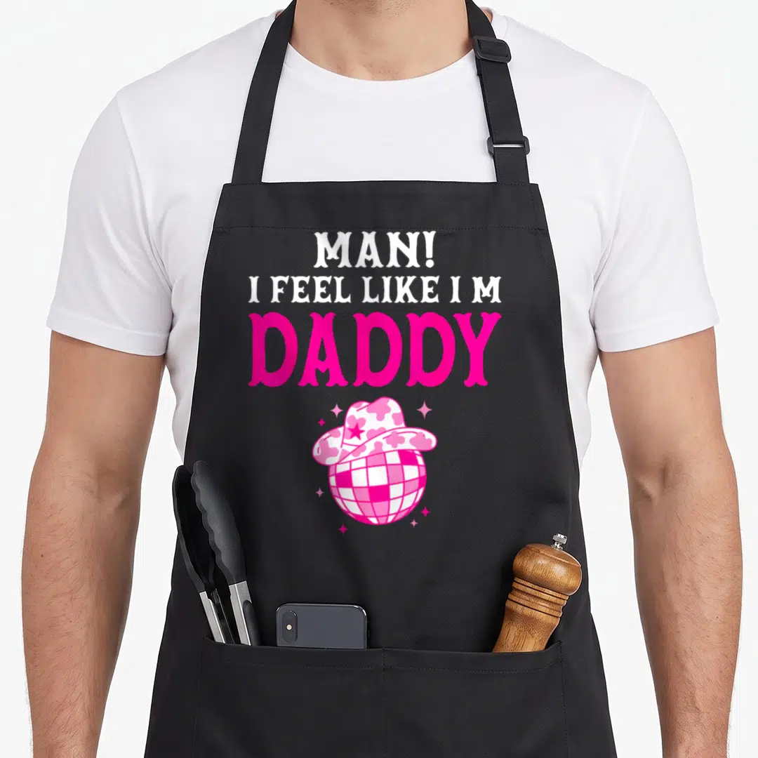 Feel cowgirl aprons - Pink disco ball wearing cowboy hat with stacked MAN I FEEL LIKE I M DADDY text — shown on a black cotton canvas apron against a white background